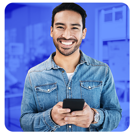 Homem sorrindo, de camisa jeans, segurando um celular, com fundo azul em ambiente de escritório desfocado.