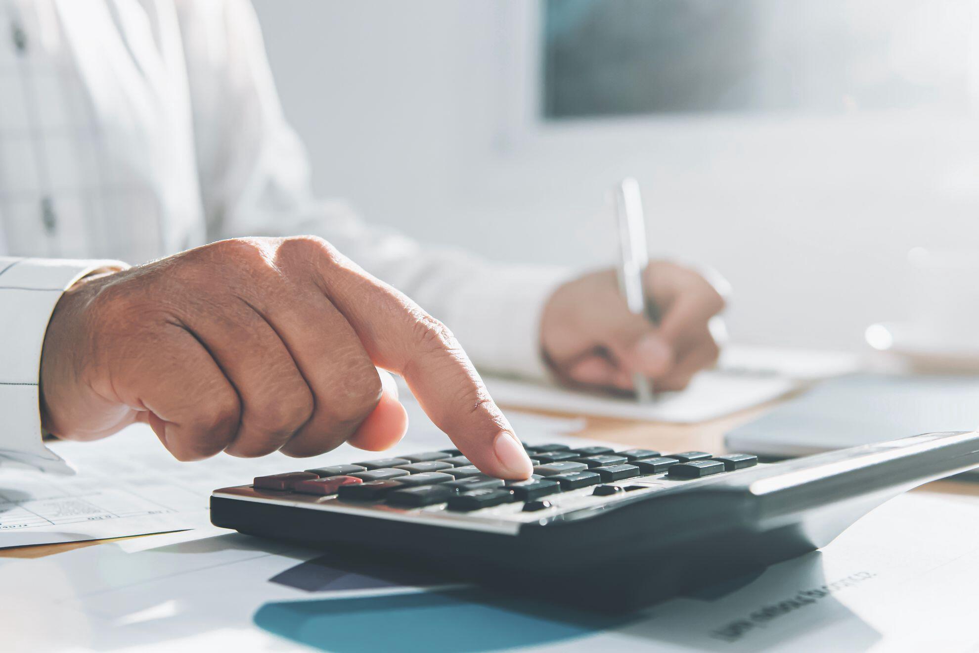 mãos masculinas digitando em uma calculadora e fazendo anotações em um papel em uma mesa de escritório.