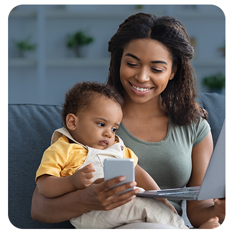 Mãe jovem, negra, sorrindo, com um bebê negro no colo, sentada no sofá de casa, mexendo no celular e segurando um notebook sobre um fundo cinza azulado.