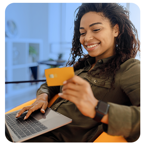 Mulher jovem, preta, sorrindo, sentada em uma poltrona laranja, com um notebook no colo, segurando um cartão de crédito laranja.