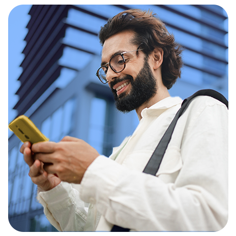 Homem jovem, branco, sorrindo, de casaco branco, segurando o celular e olhando para ele com prédios atrás dele.