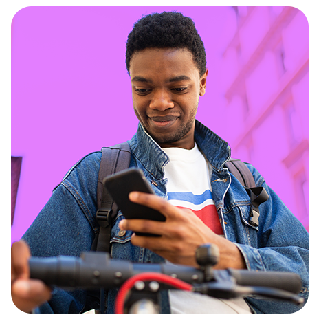 Jovem preto de jaqueta jeans, camiseta branca, segurando uma bicicleta pelo guidão, enquanto olha o celular, sobre um fundo lilás.