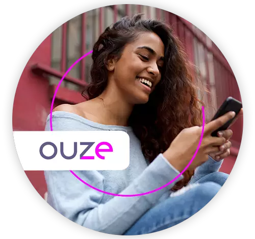 Mulher jovem, negra, sorrindo, sentada do lado de fora do prédio onde mora, olhando o celular 